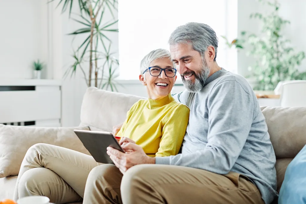 An older couple, man and wife, sitting on the couch in their living room using a tablet wirelessly connected to a Bluetooth hearing aid to facetime with a friend